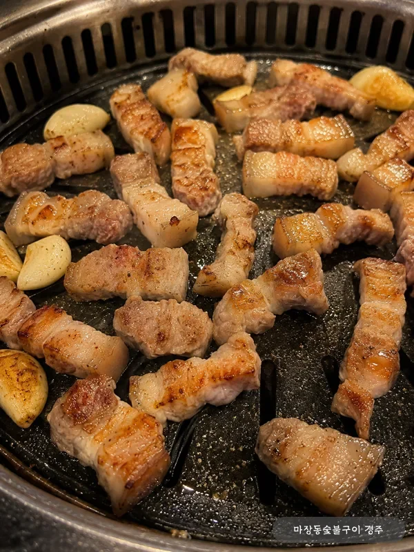 불판 가득 노릇하게 익어가는 돼지 오겹살과 통마늘 오버헤드 컷 — 컷팅 후 단면이 익어가는 단계