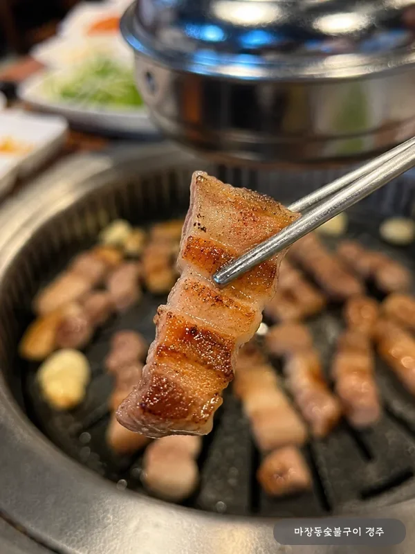 젓가락으로 들어올린 한 점의 노릇하게 구워진 돼지 오겹살 — 두꺼운 비계와 살코기 단면 클로즈업